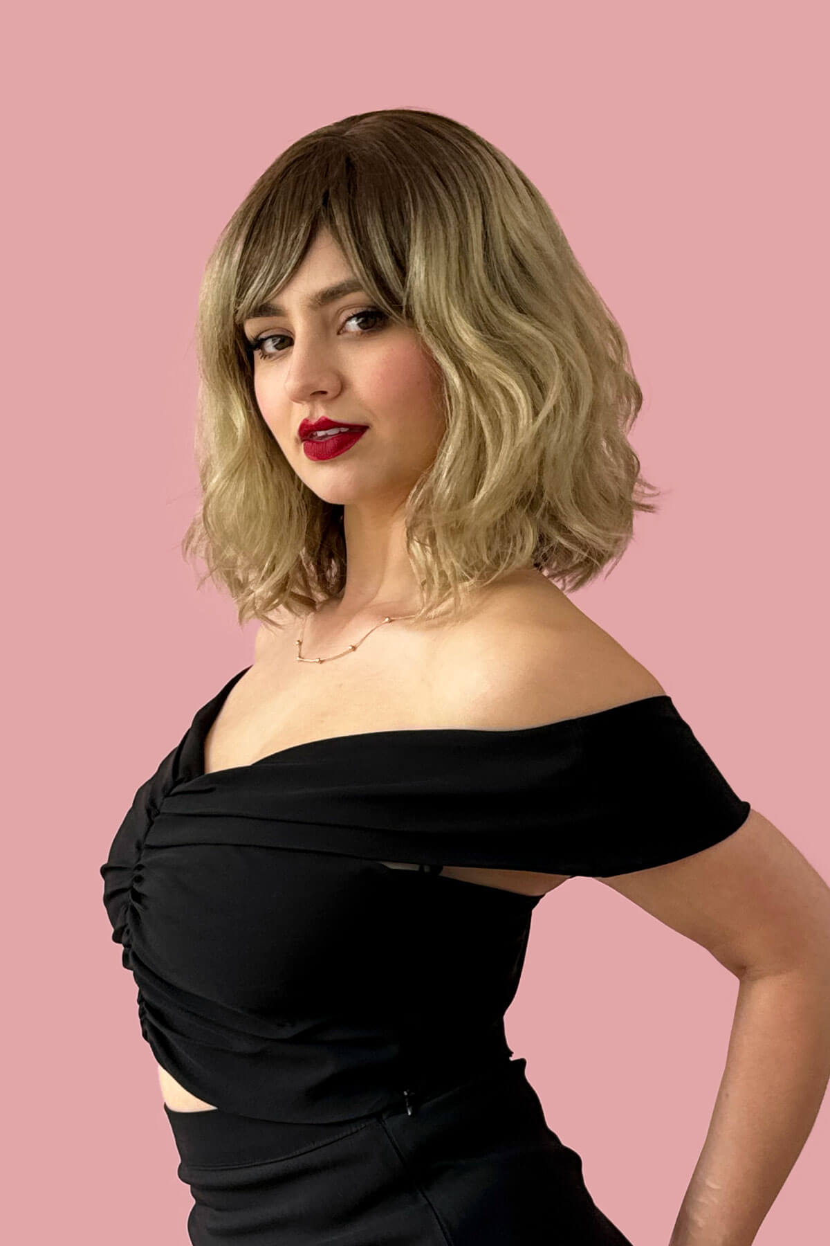 Woman with a stylish haircut and red lipstick wearing a black off-shoulder top against a pink background. Long wavy bob (lob) wig, ombre - light brown blending to ash blonde: Bianca