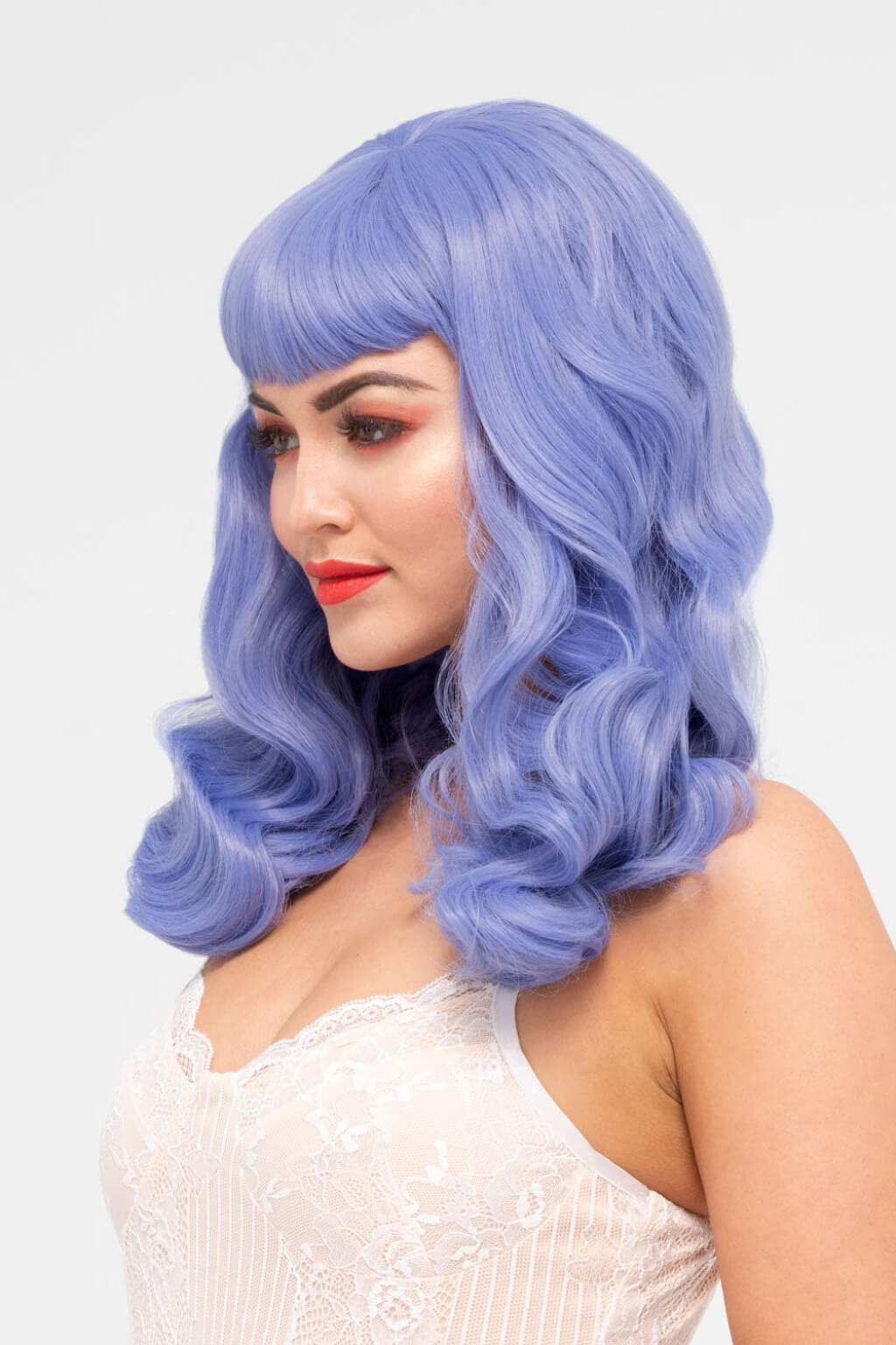 Woman with a blue wig wearing a white lace top on a light grey background. Long blue-purple wig with short fringe and old Hollywood waves: Ophelia