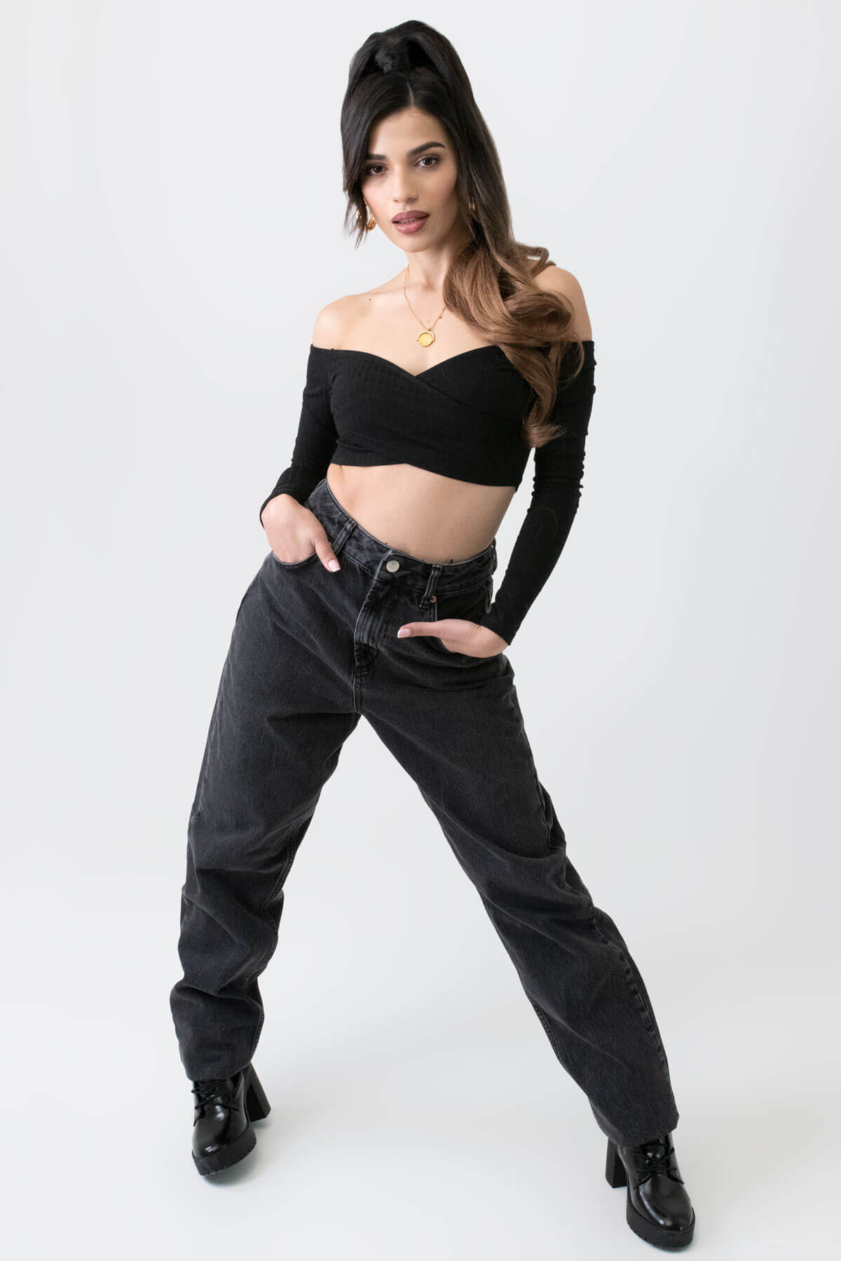 Woman wearing a black crop top and high-waisted black jeans on a white background. Long ponytail extension with soft waves: Sophie brown to blonde ombre