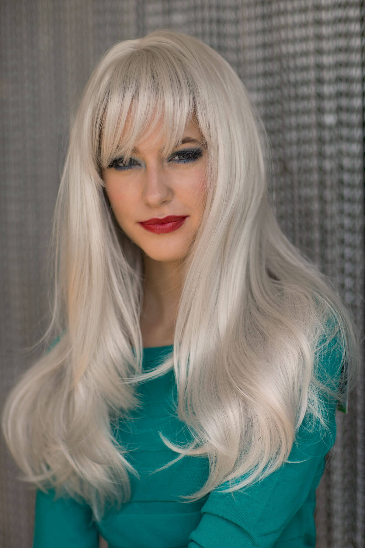 Person with long blonde wig and teal top against a textured grey background. Long silver/grey wig in razor cut, face framing style: London