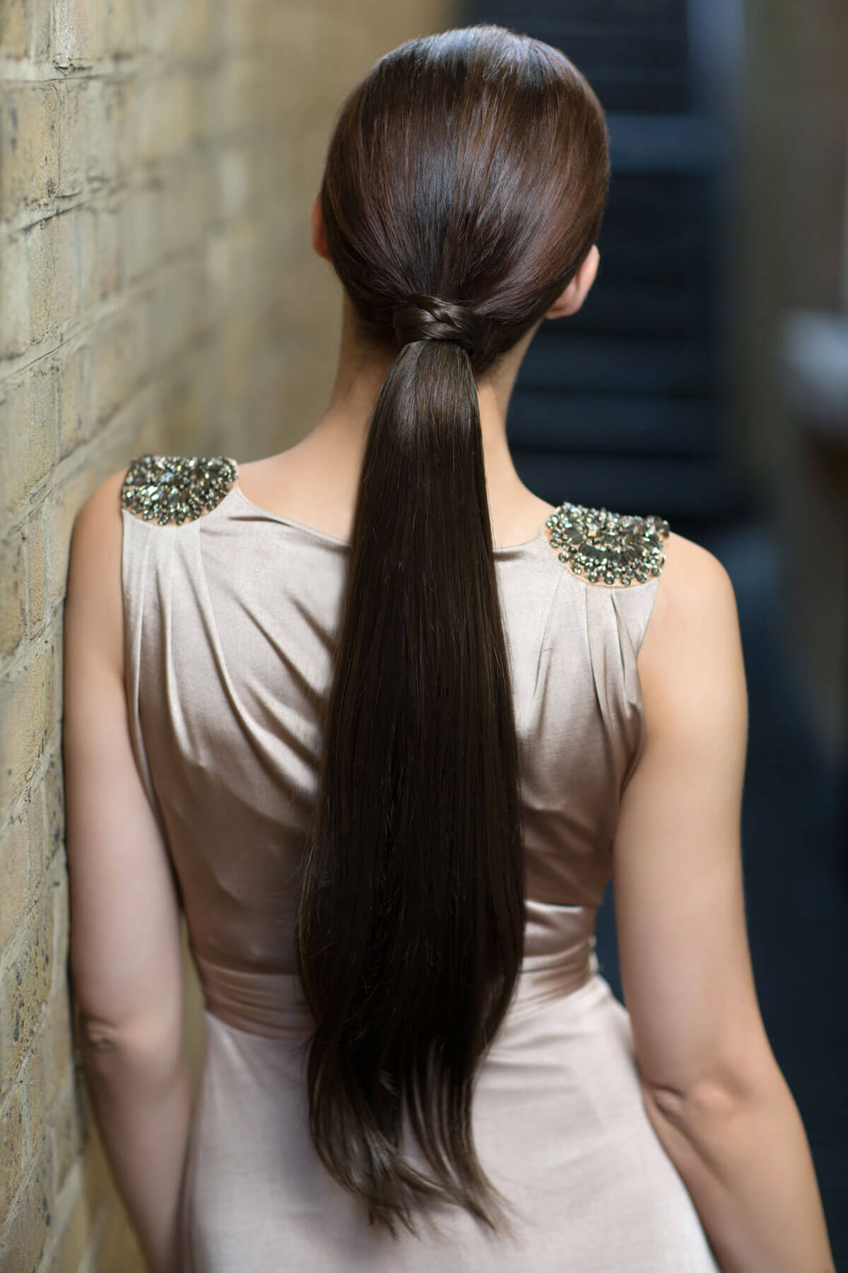 Woman with long dark hair styled in a high ponytail, wearing a light-colored dress with decorative shoulder embellishments.