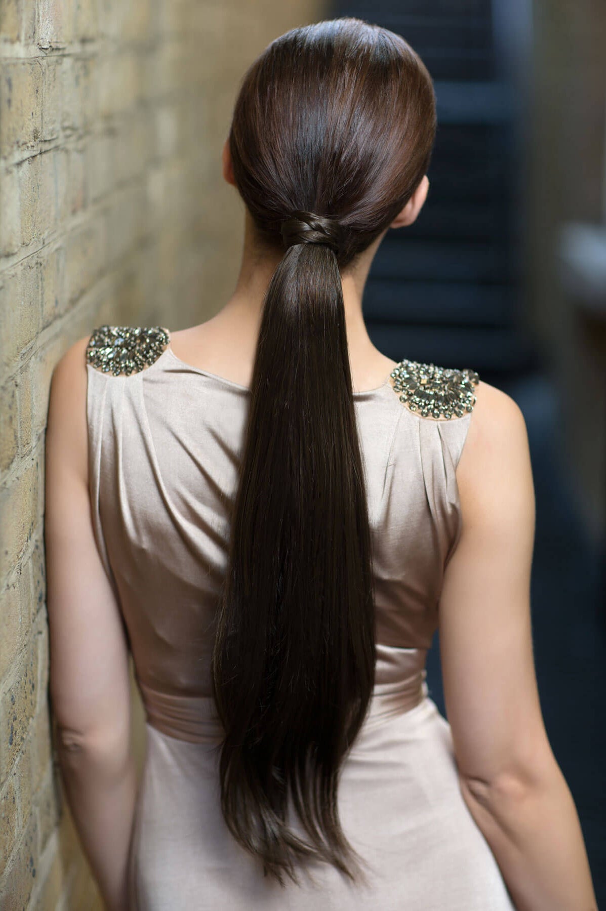 Woman with long dark hair styled in a high ponytail, wearing a light-colored dress with decorative shoulder embellishments. Long sleek ponytail extension with gentle, layered waves: Mali chestnut brown 8