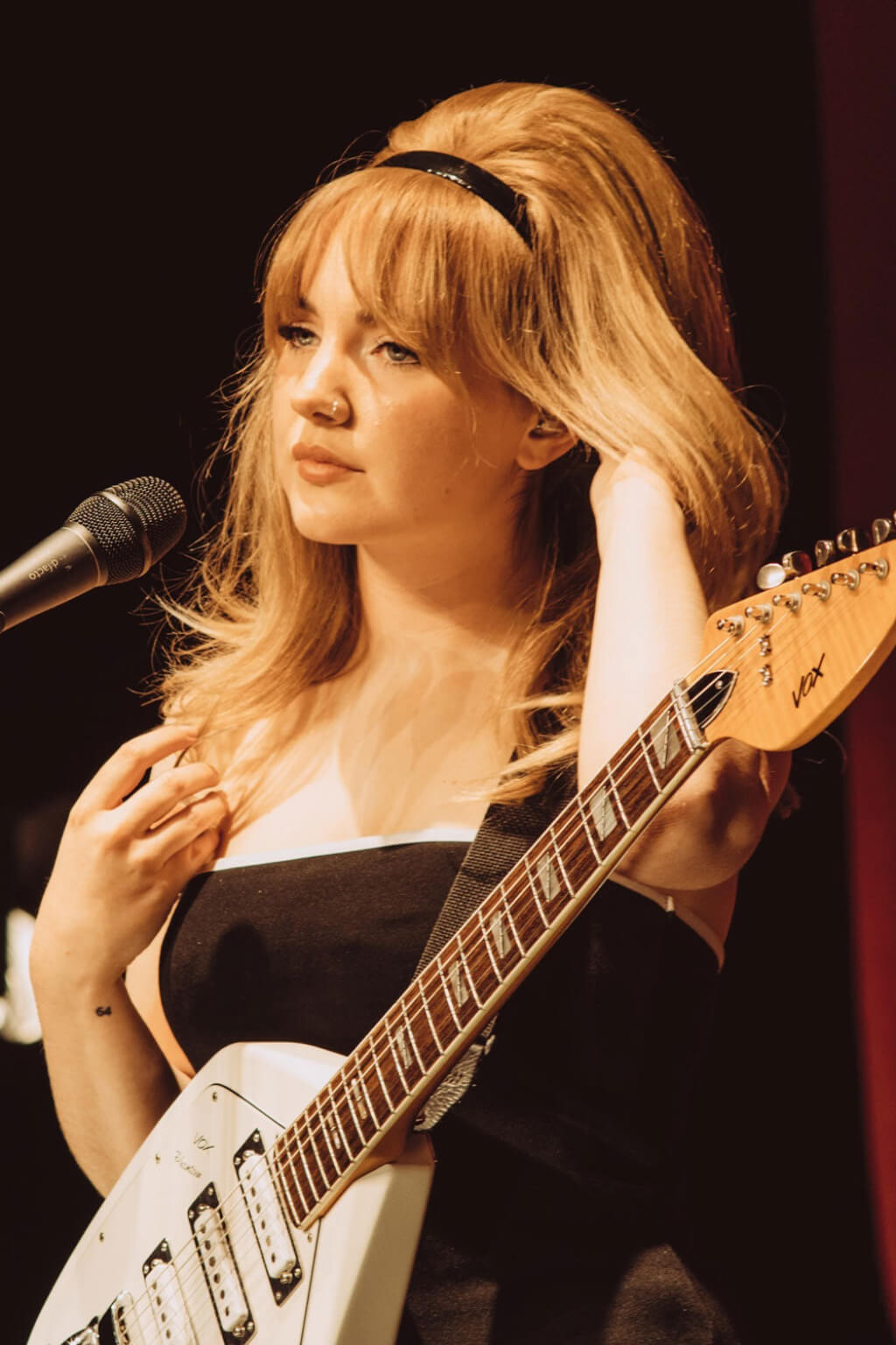 Woman with blonde hair playing a bass guitar in front of a microphone with a dark background. Volume and Height Booster Hairpiece: Blaise blonde 24H613