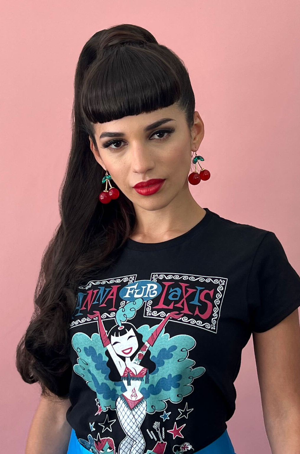 Woman wearing a black t-shirt with colorful graphics and text against a pink background. Clip-in fringe hairpiece/clip-in bangs, thick, straight retro rockabilly style: Ida dark brown 4