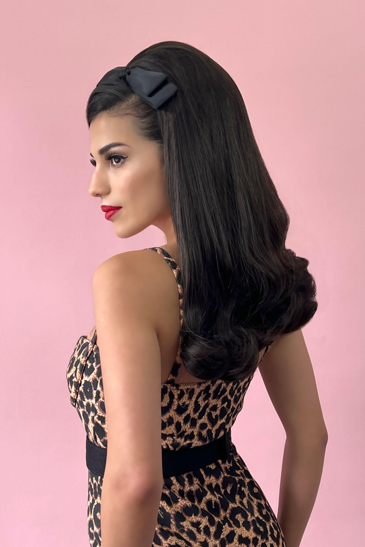 Woman with long, wavy hair wearing a black headband against a pink background. 1960s Retro high crown, half wig hairpiece, long with hair flip: Priscilla platinum blonde 1001