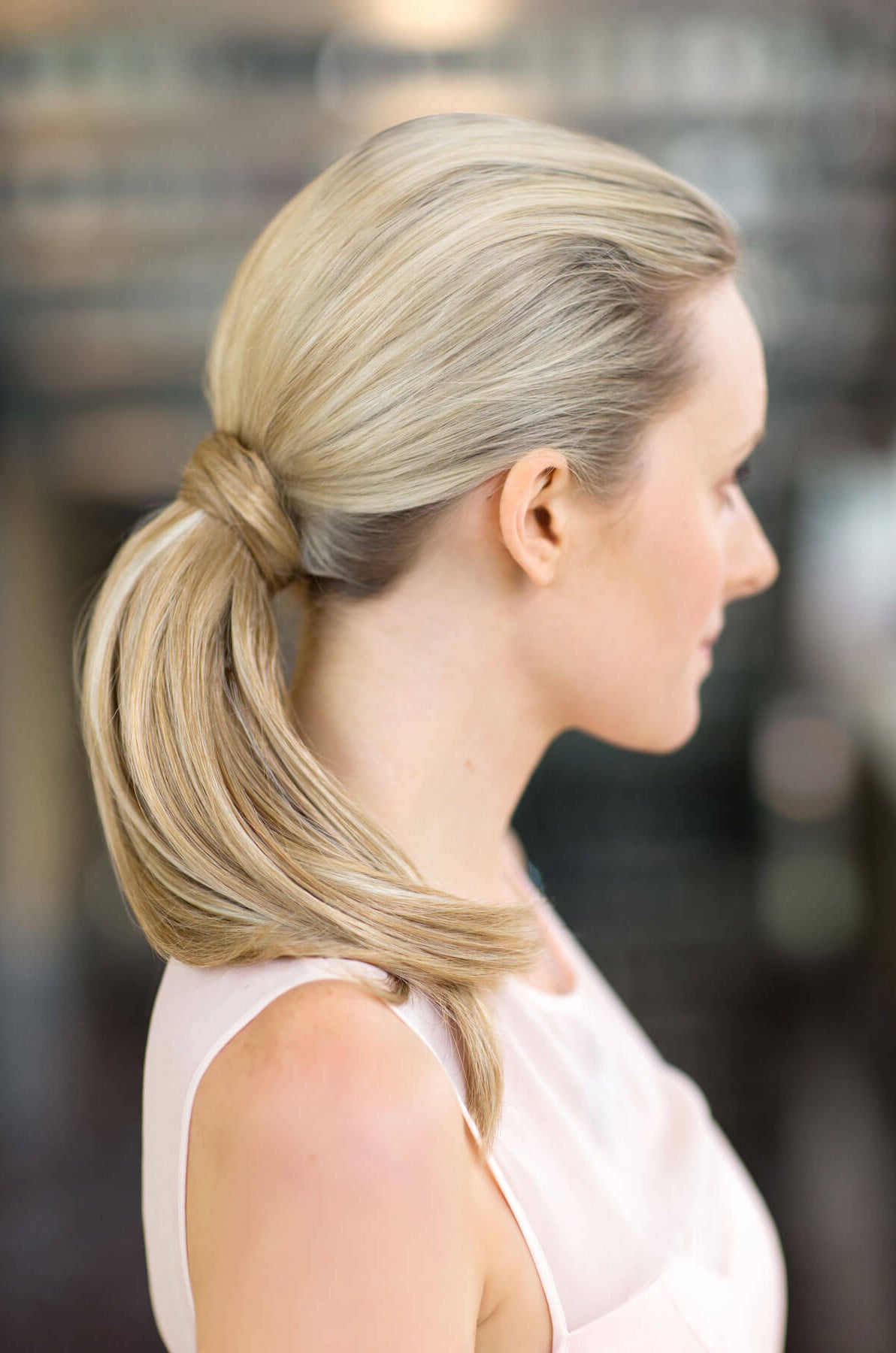 Woman with blonde hair styled in a ponytail against a blurred background. Sleek ponytail extension hairpiece, Flexihair: Jasmin blonde 24H613
