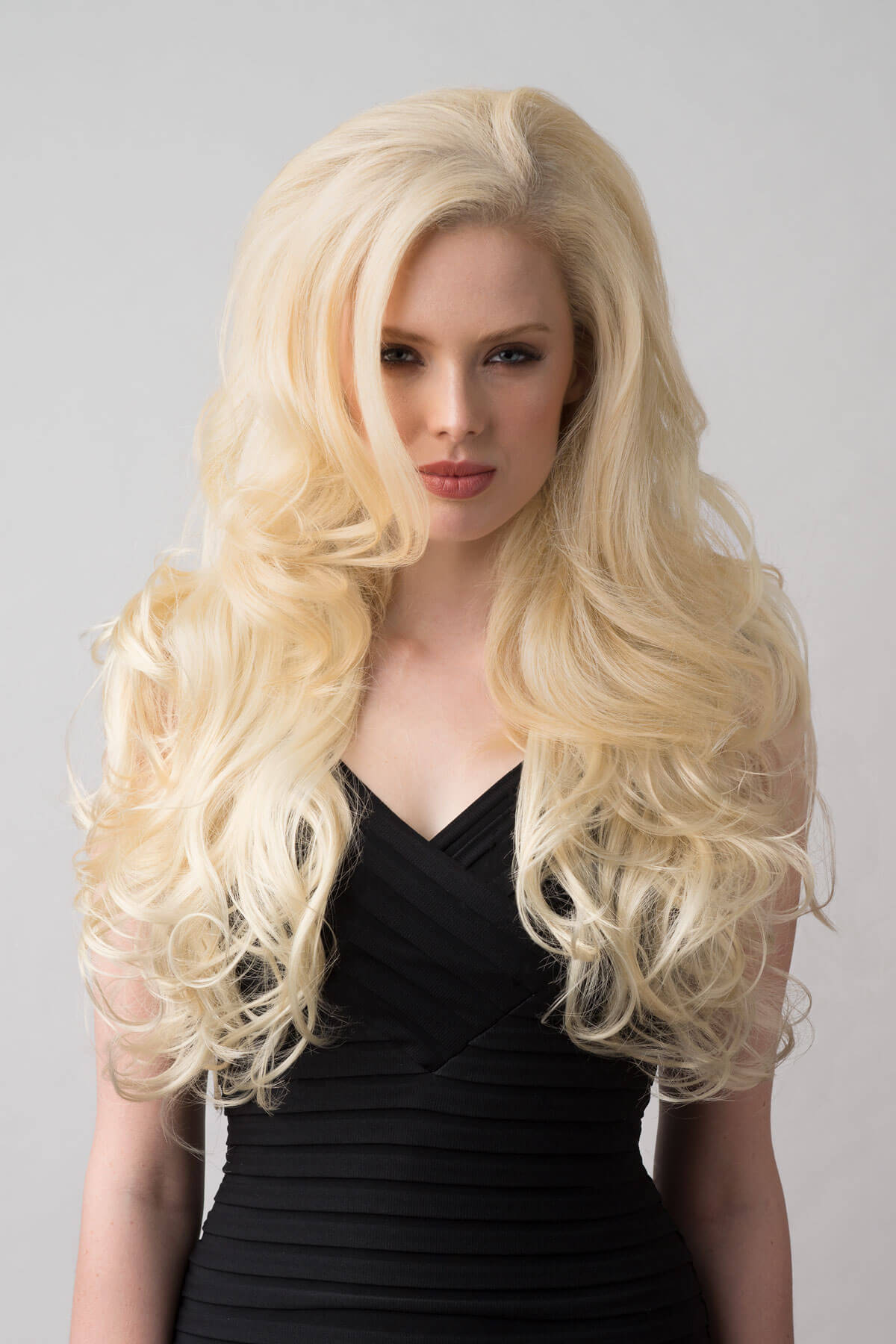 Woman with long blonde wig wearing a black dress on a gray background. Long blonde 3/4 hairpiece (half wig), big loose curls: Zara creamy blonde 613