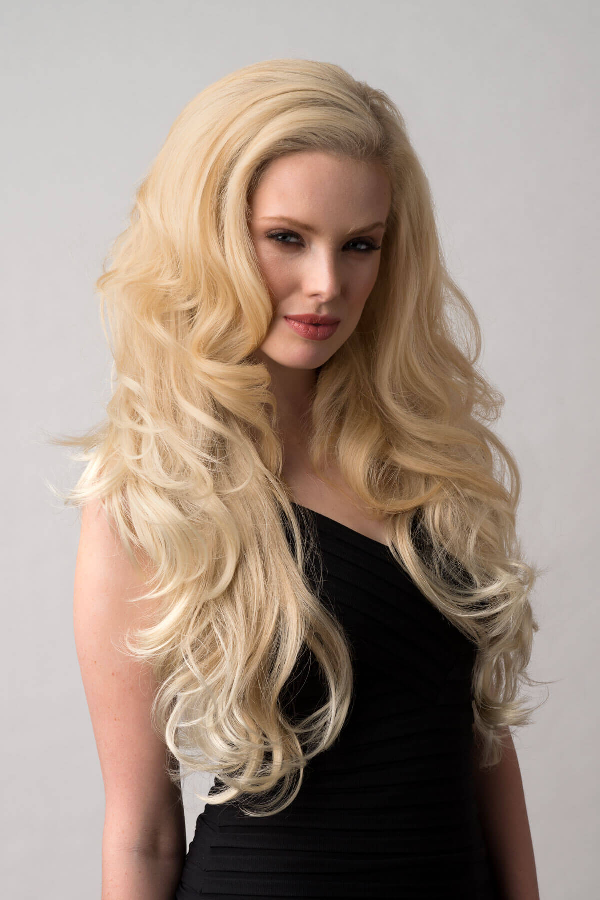 Woman with long blonde hair wearing a black dress on a gray background. Long blonde 3/4 hairpiece (half wig), big loose curls: Zara blonde 613T24