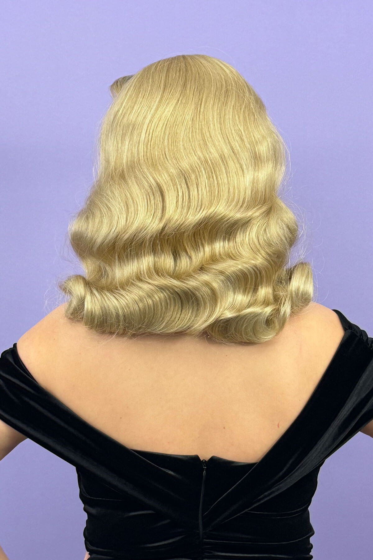 Blonde wig with wavy hair on a person wearing a black dress against a purple background. 1950s style half wig hairpiece with deep finger waves: Deborah blonde 613T24