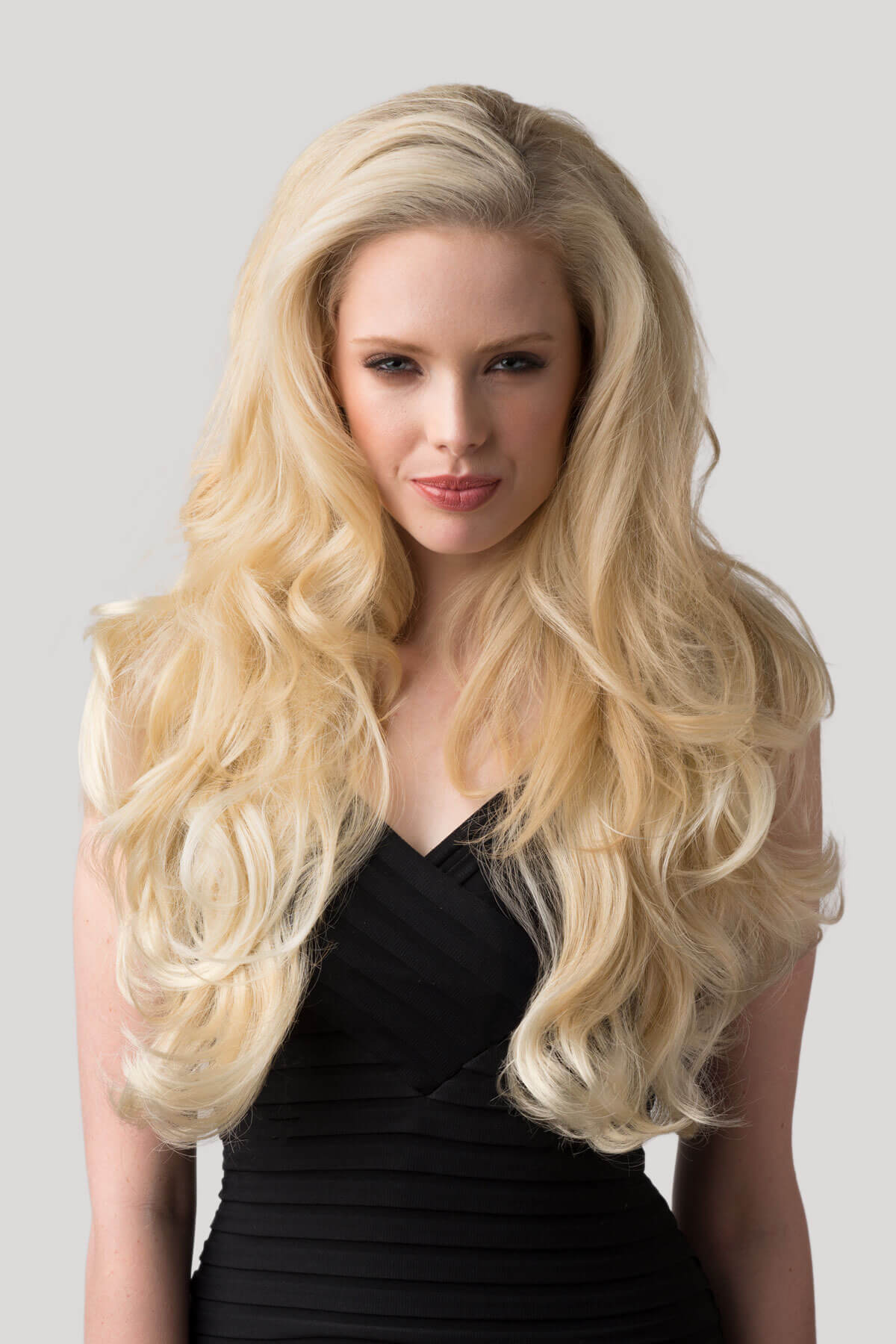 Woman with long blonde hair wearing a black dress on a light gray background. Long blonde 3/4 hairpiece (half wig), big loose curls: Zara blonde 24BT613