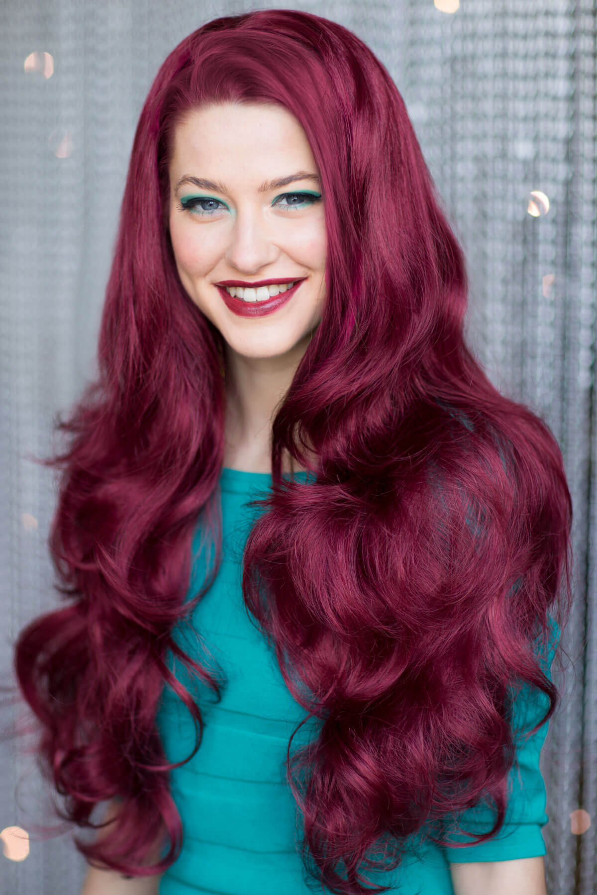 Woman with vibrant red hair and a teal top against a textured gray background. Red half wig hairpiece (3/4 wig), big loose curls: Verity wine red 39