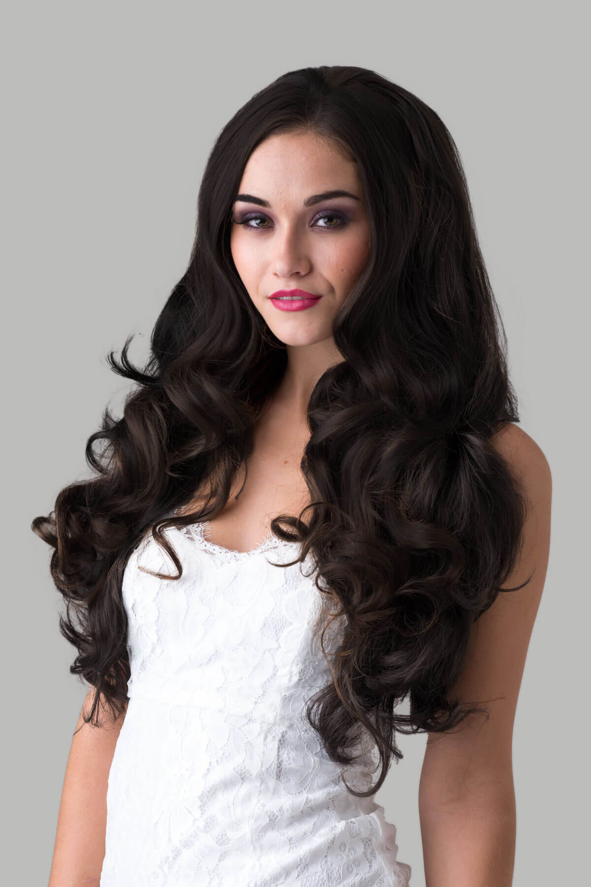 Woman with long, wavy hair wearing a white top against a gray background.  Brown half wig hairpiece with long loose curls: Chelsea dark brown 4