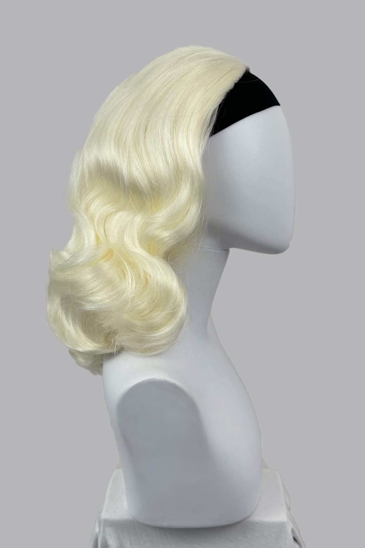 Blonde wig on a mannequin head against a grey background viewed from the side. 1950s style shoulder length half wig hairpiece with beautiful vintage waves: Onyx platinum blonde 1001