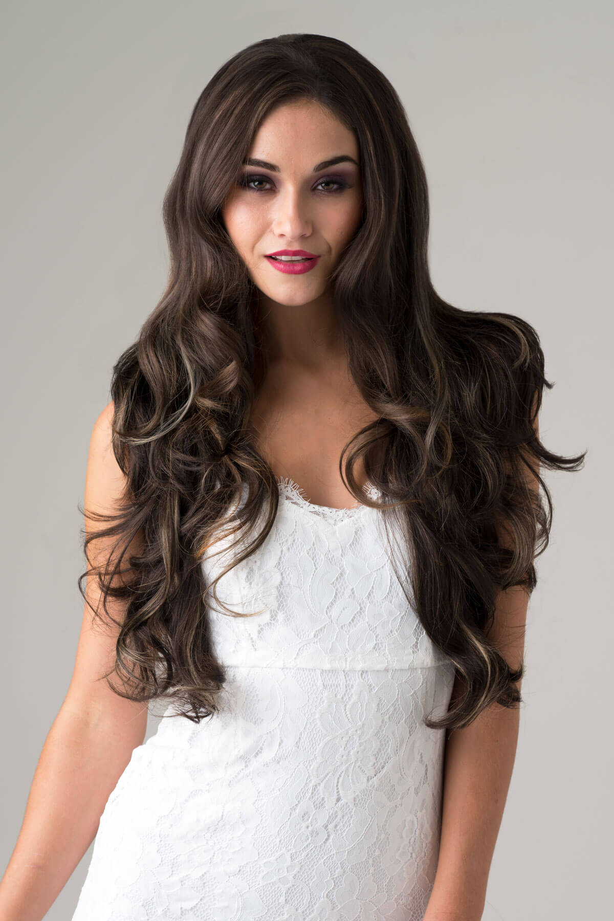 Woman with long, wavy brown hair wearing a white dress against a gray background. Brown half wig with blonde highlights: Reese