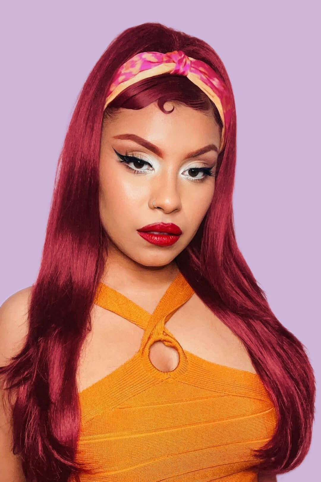 Woman with vibrant red hair and orange top against a light purple background. 1960s style hair, high crown long straight and layered half wig: Juliette wine red 39