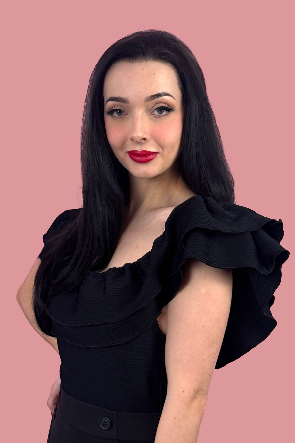 Woman with long black hair wearing a black off-shoulder top with ruffled sleeves against a pink background. Straight black hairpiece (3/4 wig), long and layered: Raven