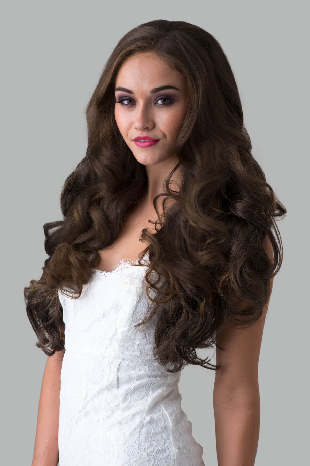 Woman with long, wavy brown hair wearing a white dress against a gray background. Brown half wig hairpiece with long loose curls: Chelsea chestnut brown 8
