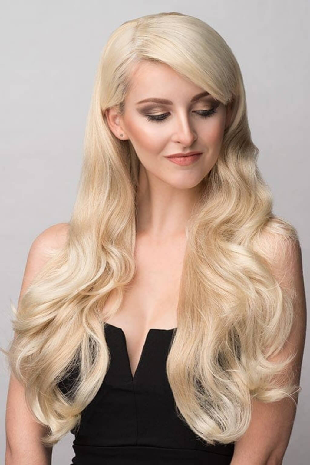 A woman with long, wavy platinum blonde hair wearing a black strapless top, shown from the chest up against a plain light grey studio background. Blonde half wig hairpiece (3/4 wig), loose curls: Suki blonde 613T24