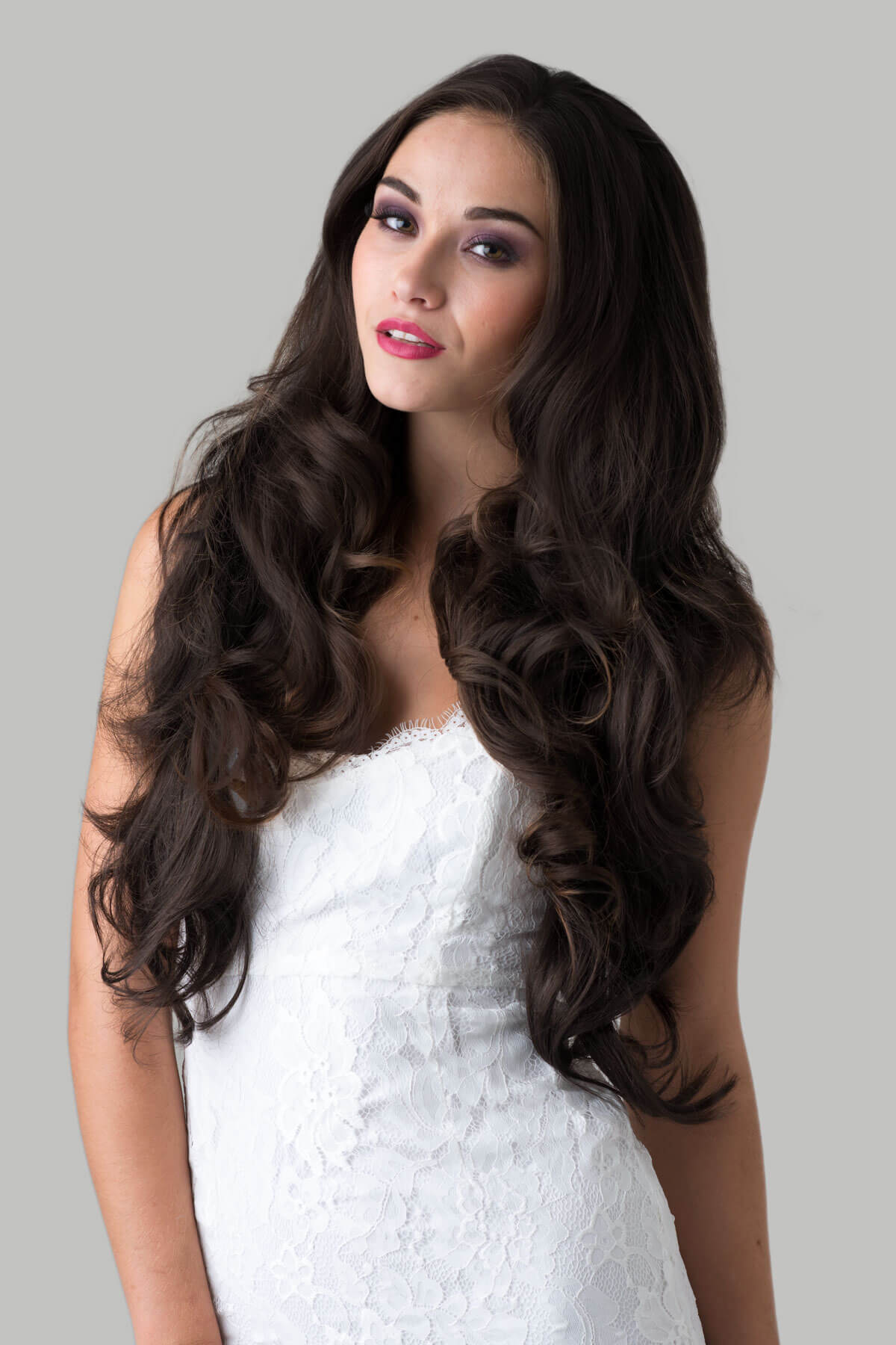 Woman with long, wavy brown hair wearing a white dress against a gray background. Brown half wig hairpiece, full, long with gentle waves: Taya dark chocolate brown 6
