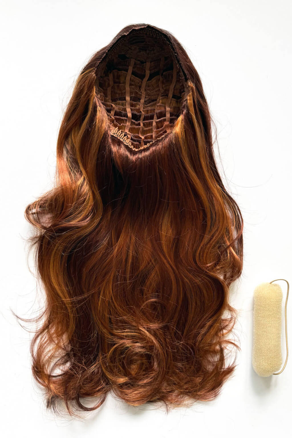 Long wavy brown wig with warm auburn highlights, shown from the top with the internal lace hairline visible, alongside a small beige hair roller on a white background. High crown long, wavy half wig and volume ring: Bardot Auburn blonde BO