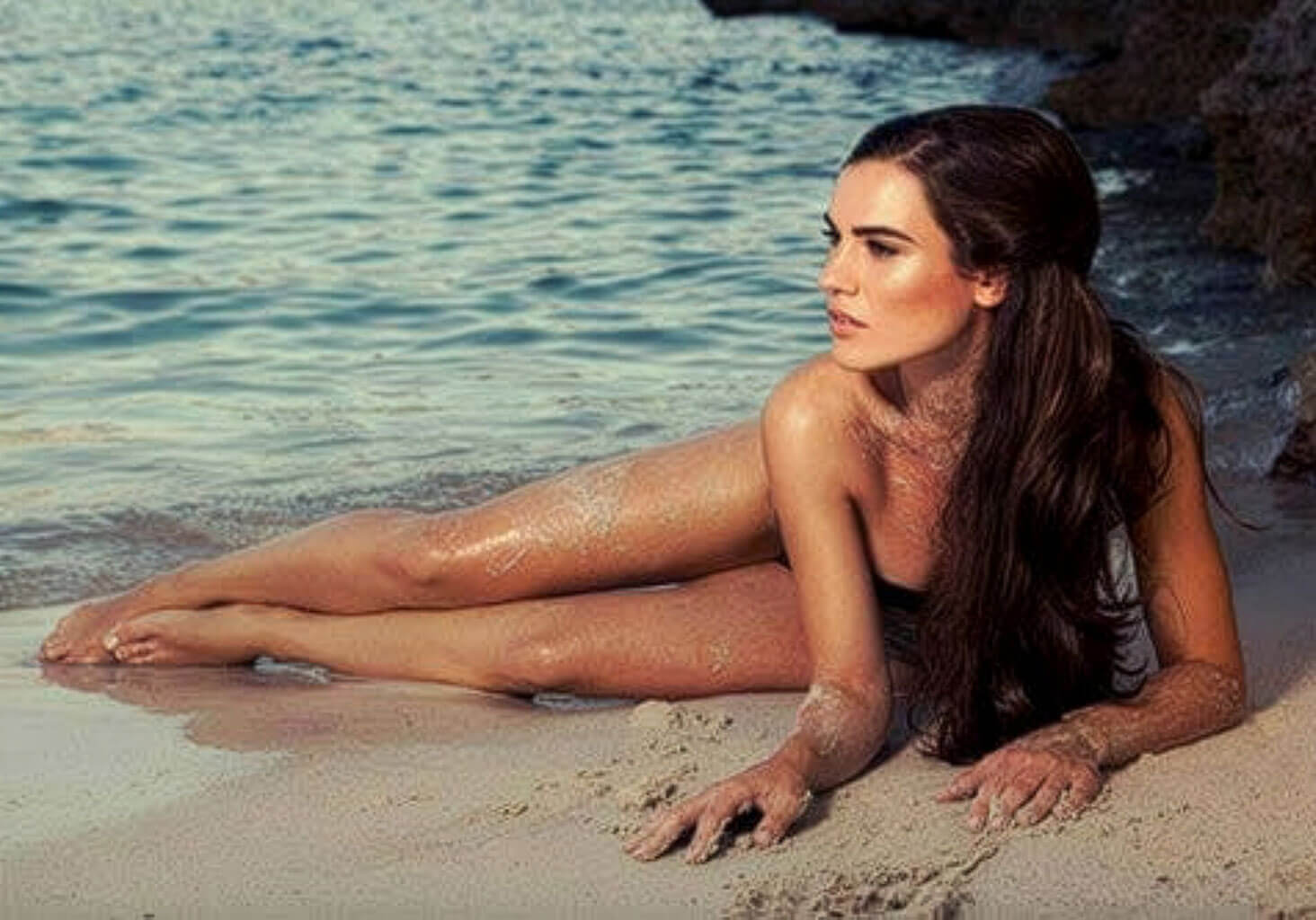 Woman lying on a sandy beach with ocean water in the background. Brown half wig hairpiece (3/4 wig), layered, wavy: Simone dark brown 6