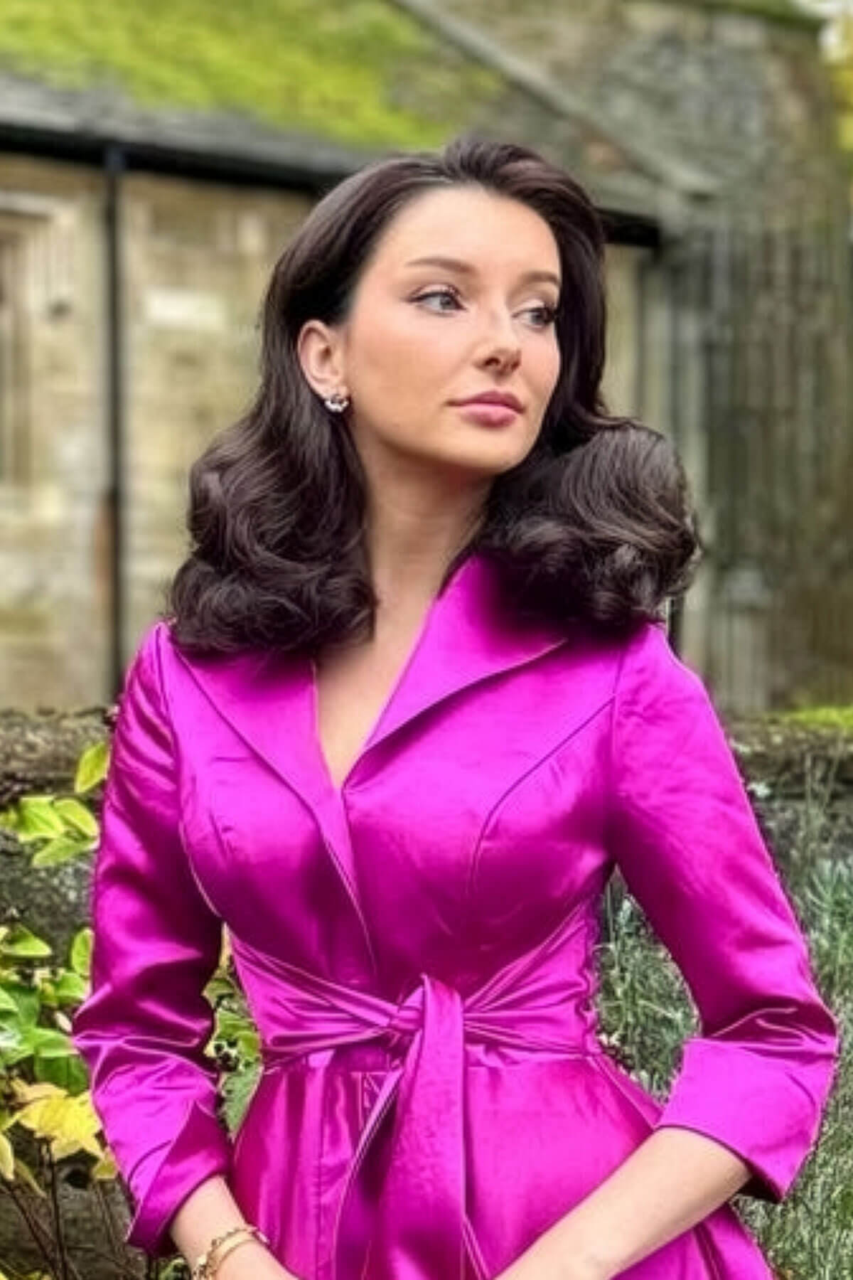 Woman in a bright pink dress standing outdoors with greenery in the background. Pinup style half wig hairpiece with beautiful marcel waves: Virginia chestnut brown 8