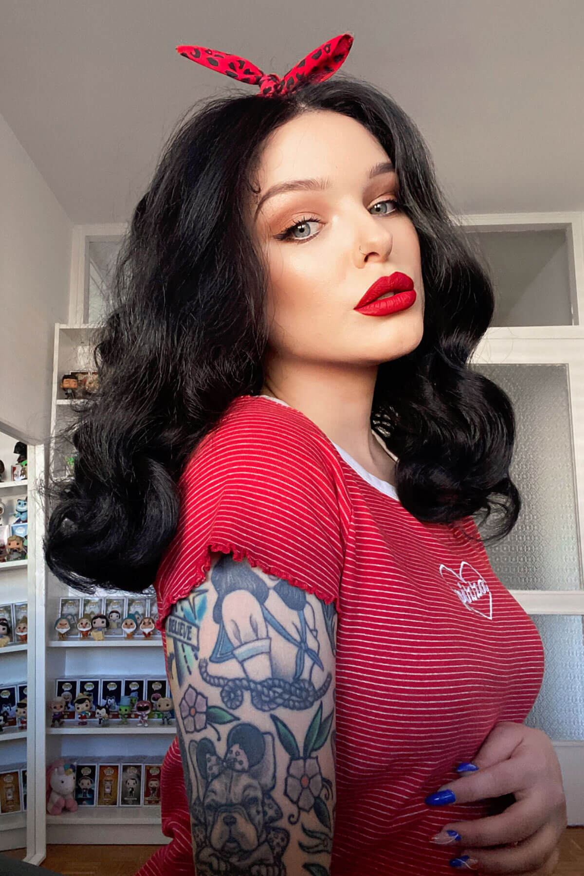 Woman wearing a red top with a heart design and black hair with a red bow indoors. Black pinup wig, lace front with long finger waves: Tyra
