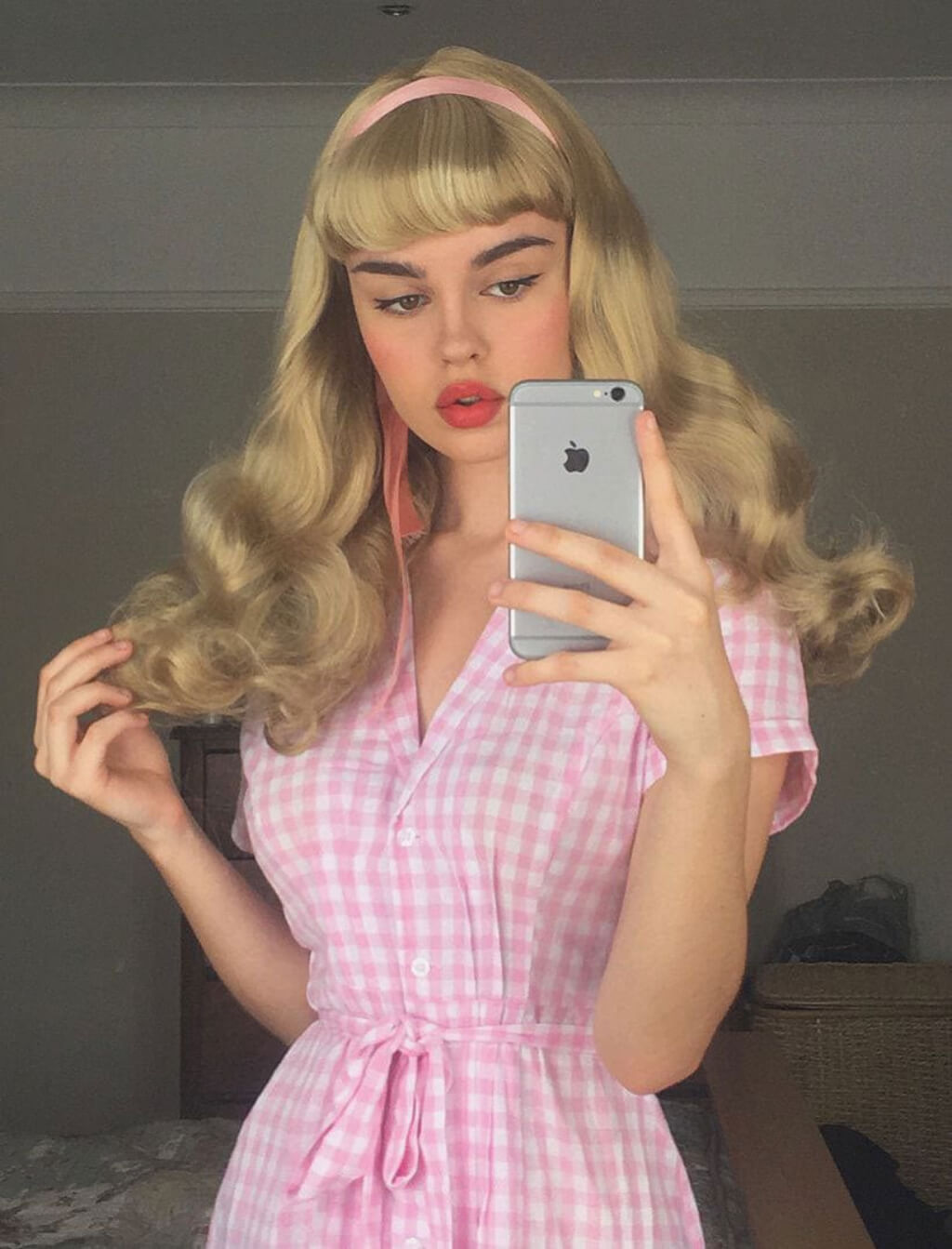 A woman taking a mirror selfie wearing a pink and white gingham dress, a pink headband, and a blonde wig with bangs. Long blonde 1950's style wig, gently wavy with short fringe: Emmeline