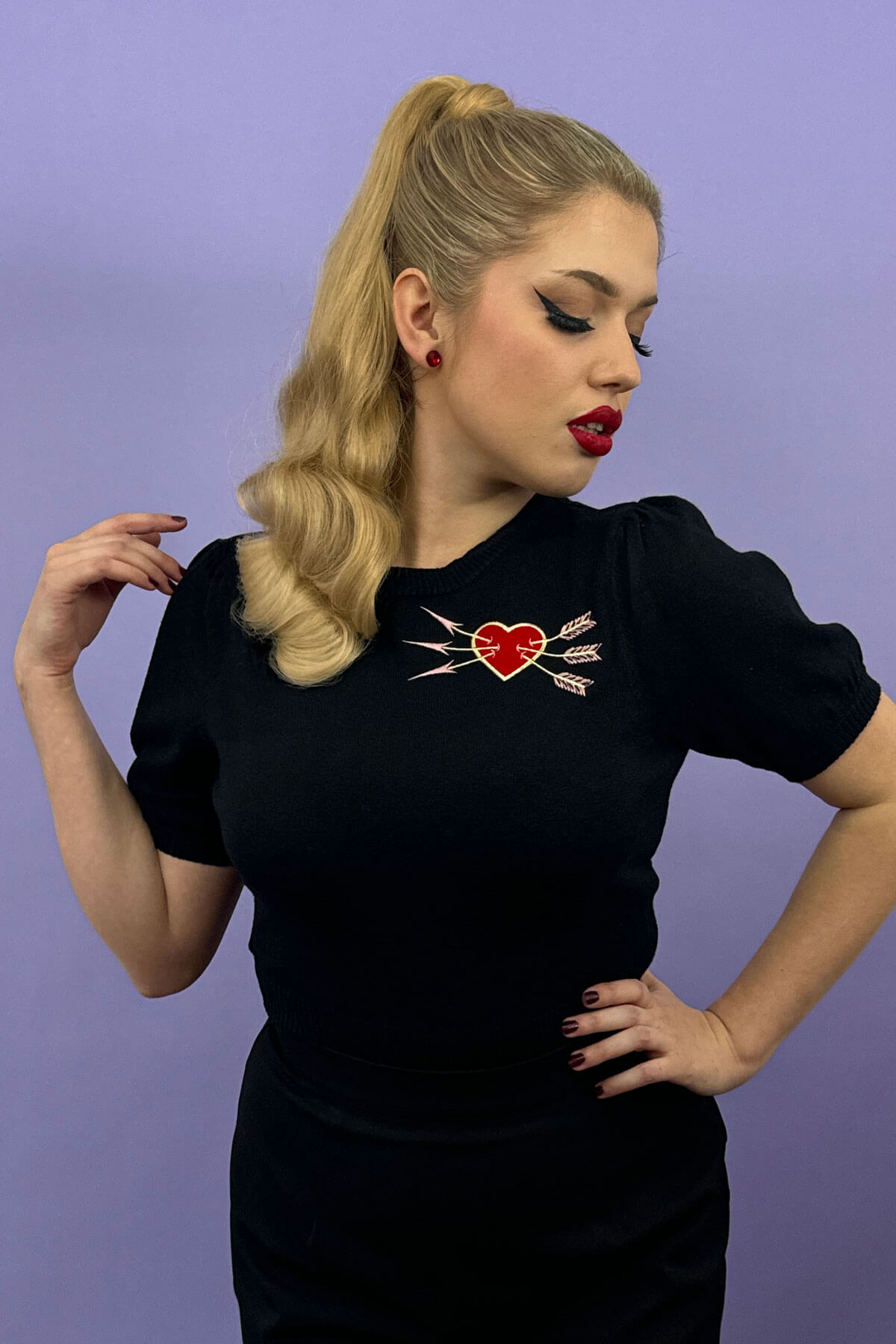 Woman wearing a black t-shirt with a red heart and white arrow design against a purple background. Retro style ponytail hairpiece, long with vintage waves: Arabella blonde 24H613