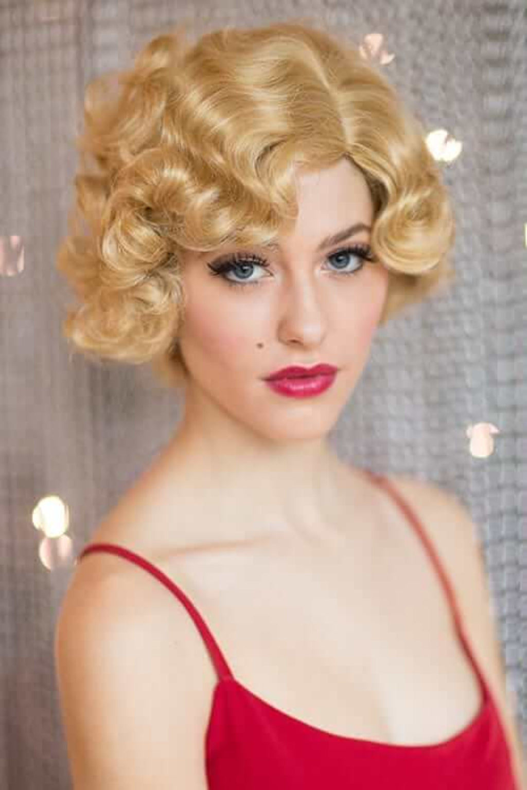 Woman with blonde curly hair and red lipstick against a blurred background. Blonde 1920s 1930s style wig, short with finger waves: Diva warm blonde 24B