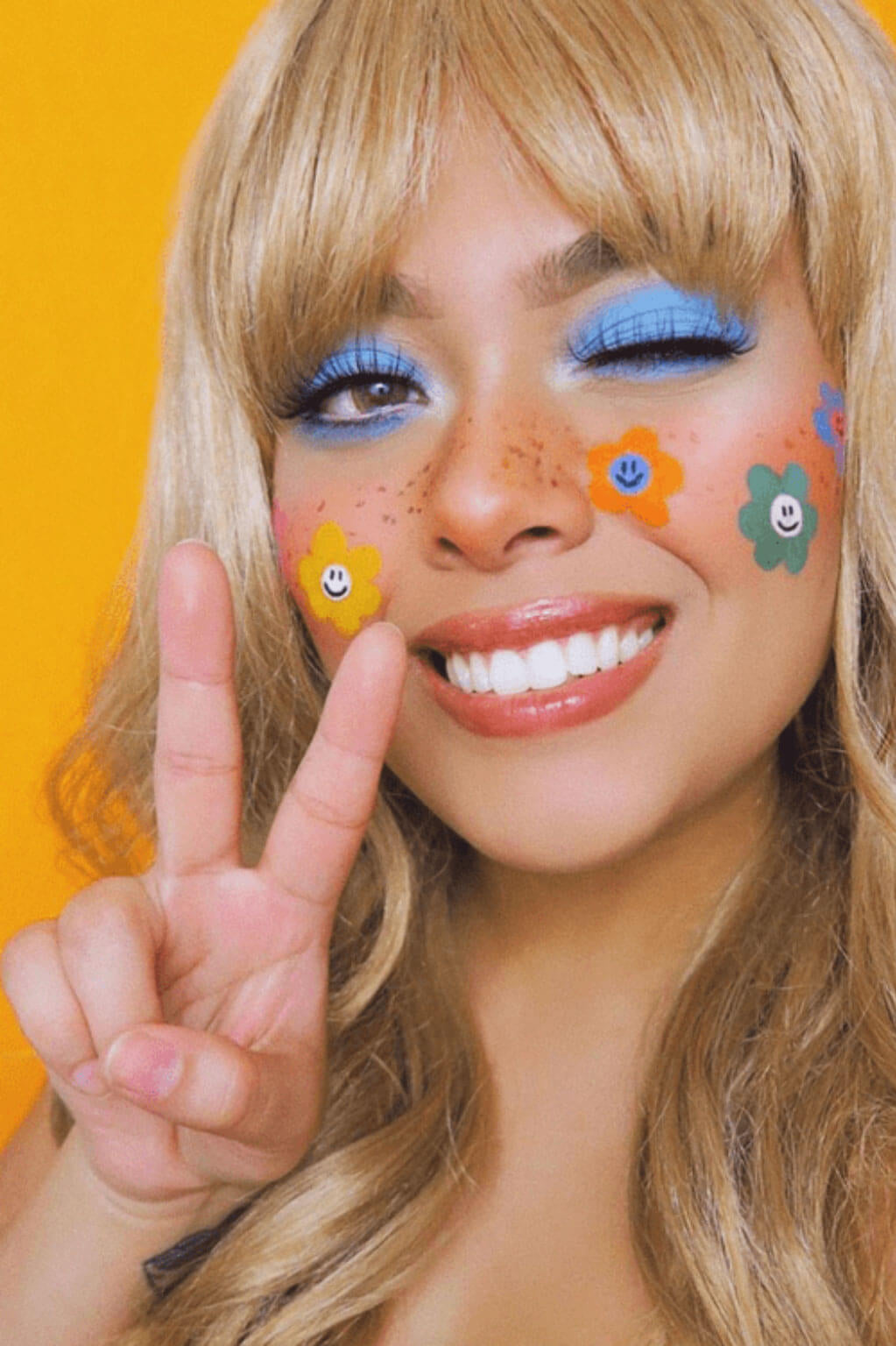 Person with colourful face paint and flowers, making a peace sign against an orange background. Lucky dip wig