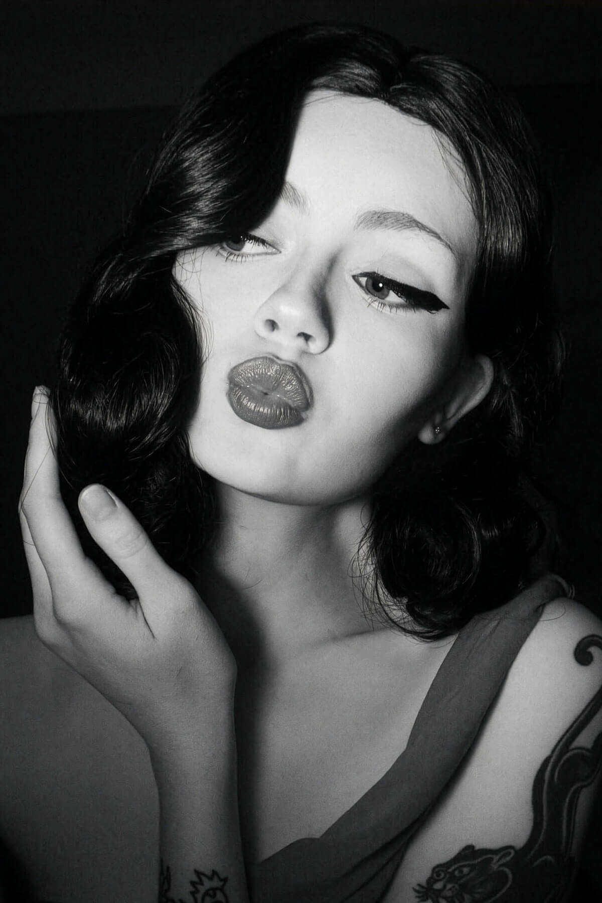 Black and white portrait of a woman with a vintage hairstyle, dark lipstick and tattoos on her arm.