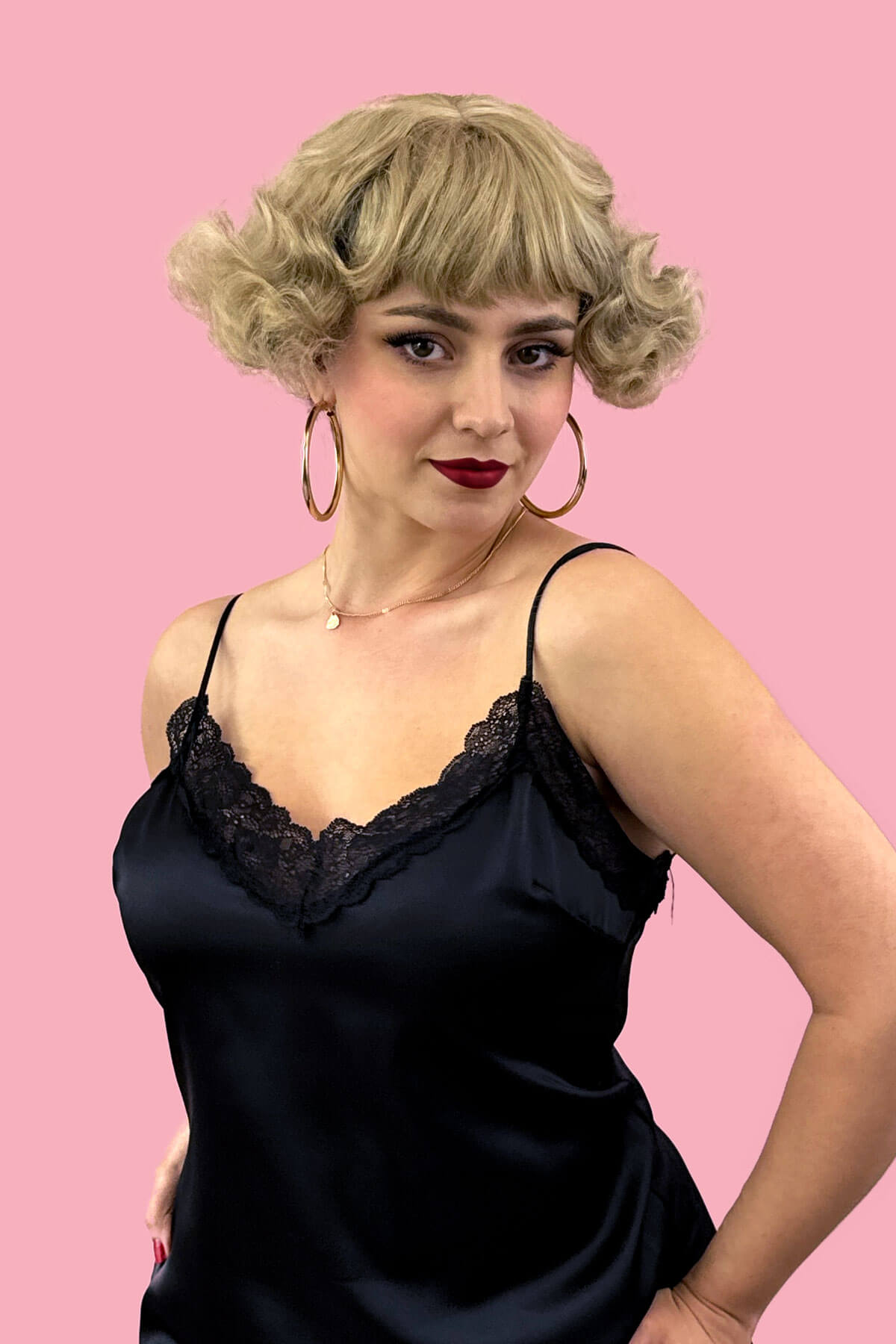 Woman wearing a black lace-trimmed dress against a pink background. A short blonde vintage style wig with waves: Joy