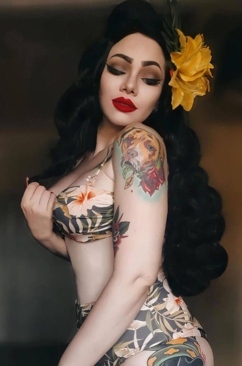 Woman with tattoos and a yellow flower in her hair against a dark background. Long black pinup style wig with vintage hollywood waves: Veronica