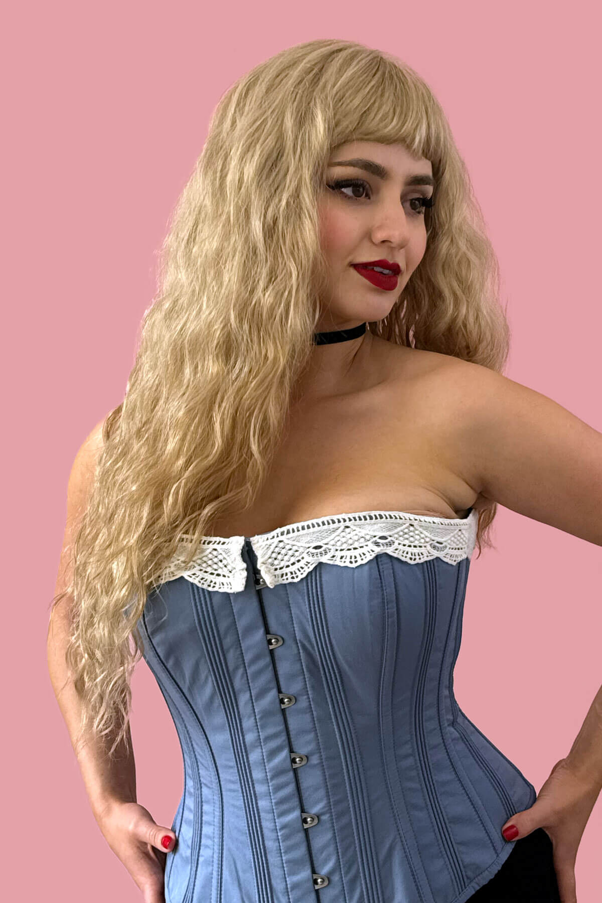 Woman wearing a blue corset with lace details against a pink background. Long wig with crimped mermaid waves: Astrid
