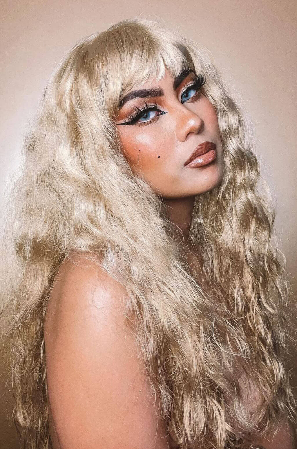 Person with long blonde hair and bold makeup against a neutral background. Woman wearing a black cap and fishnet top against a beige background. Long wig with crimped mermaid waves: Astrid