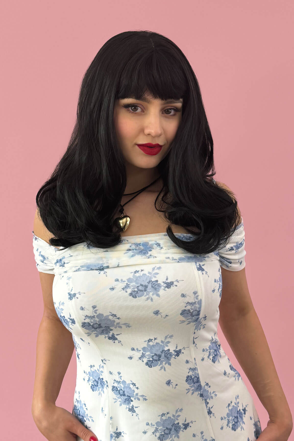 Woman with a black wig and floral dress against a pink background. Long black wavy bob (LOB) wig with straight retro fringe: Ciarra