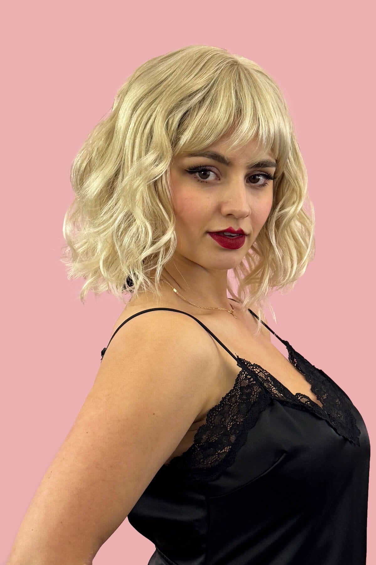 Woman with blonde wig and red lipstick wearing a black lace top against a pink background. Blonde long wavy bob (lob) wig, ash blonde blending to light blonde: Pearl blonde 613T24