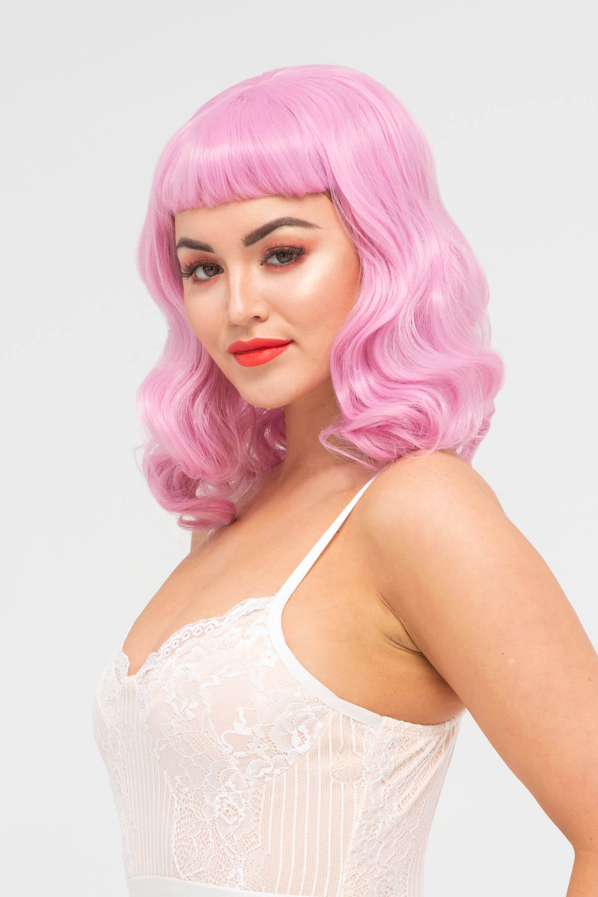 Woman with pink wig and red lipstick wearing a white lace top on a light gray background. Pink pinup style wig, with finger waves and a short fringe, 1950s style: Stevie light pink 2614