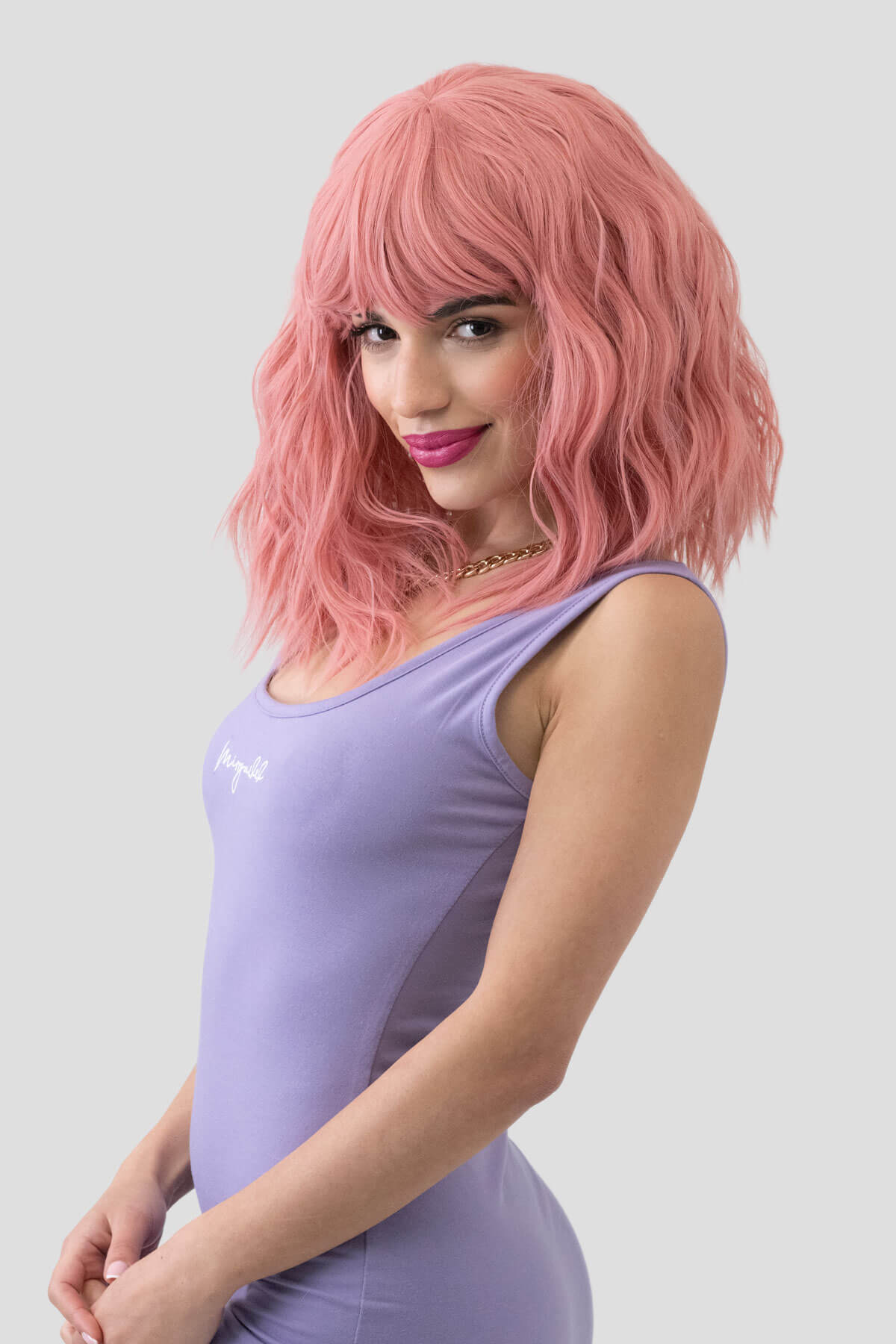 Woman with pink hair wearing a purple dress against a light gray background. Dusty pink long wavy bob wig (LOB) with textured waves: Fearne
