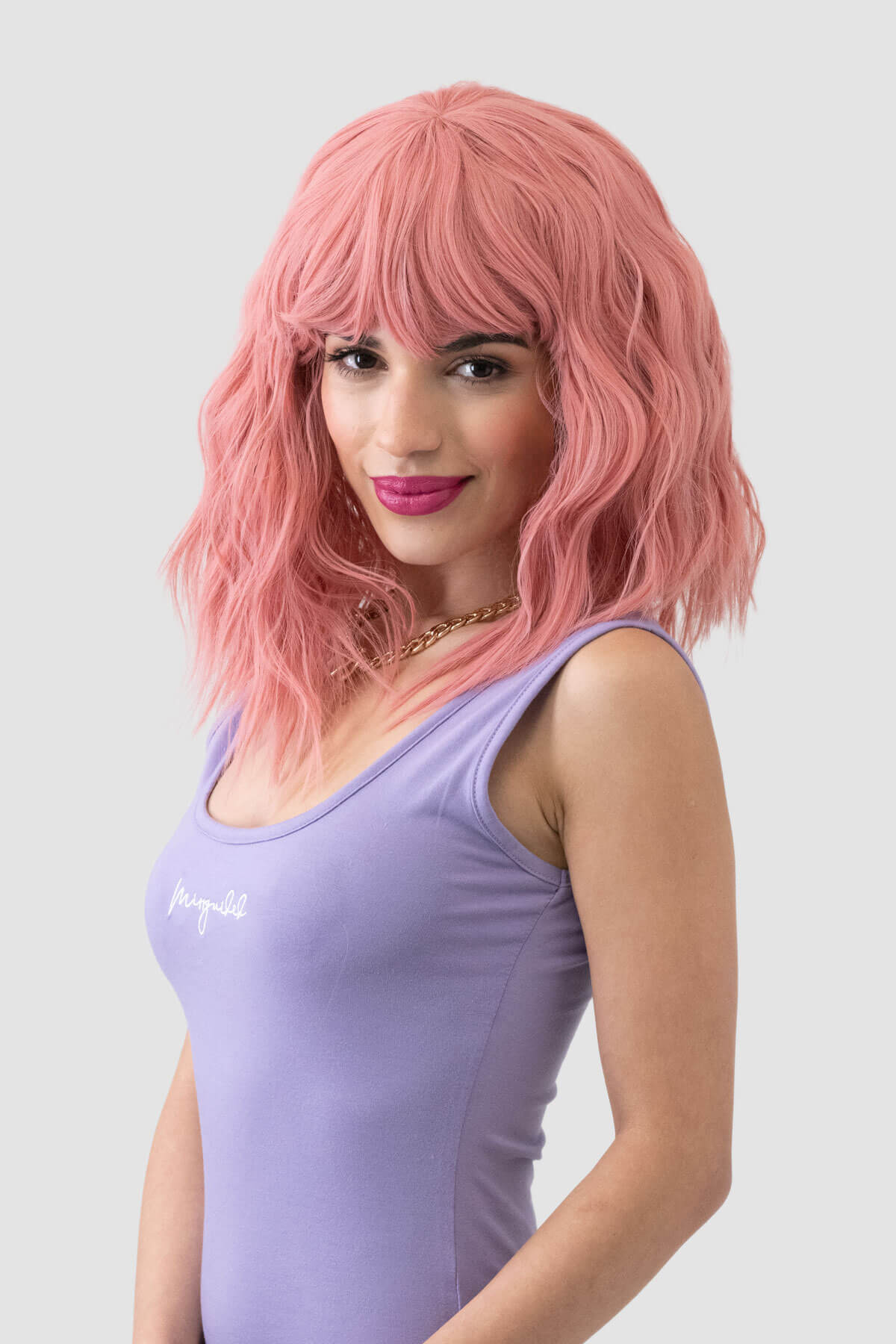 Woman with pink wig wearing a purple tank top on a white background. Dusty pink long wavy bob wig (LOB) with textured waves: Fearne
