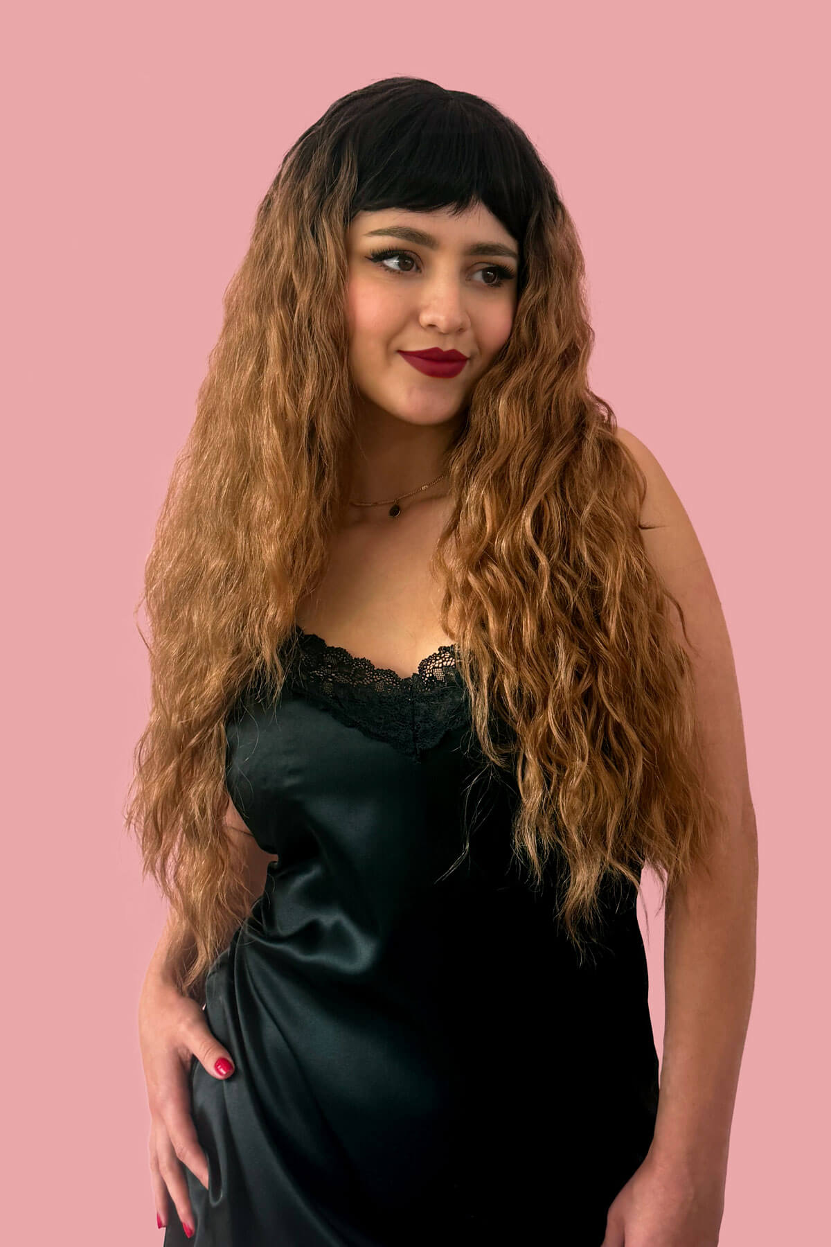 Woman with long wavy hair wearing a black dress against a pink background. Long wig with crimped mermaid waves: Astrid brown and blonde 4TN27