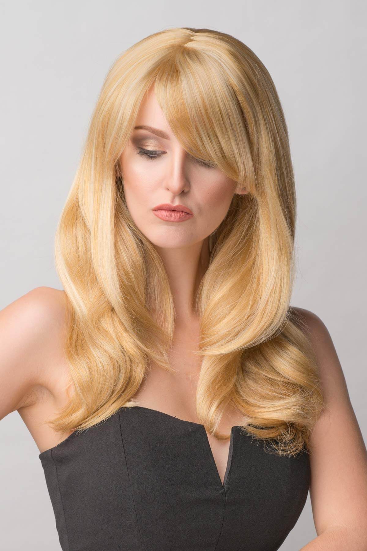 Woman with long blonde hair styled in loose waves against a gray background. Long blonde wig, face framing with long, side-sweeping fringe: Ashley honey blonde 24BH613