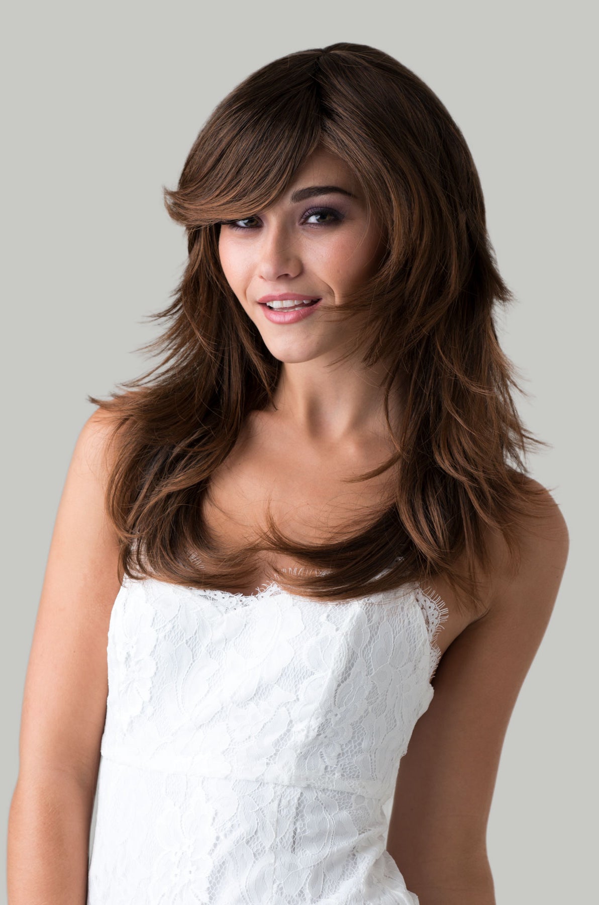 Woman with long brown hair wearing a white dress against a gray background. Long warm brown wig, layered, flicked tips: Selina