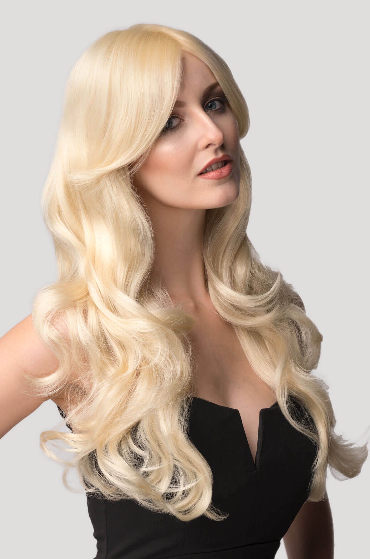Woman with long blonde wig on a plain background. Blonde wig with big loose curls and sweeping fringe, extra long: Carly champagne blonde 613