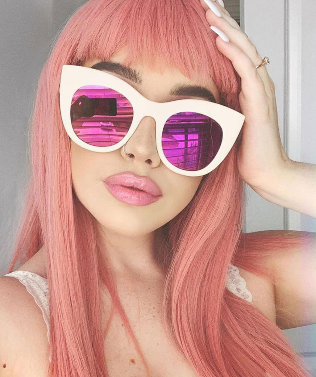 Person with pink hair wearing white sunglasses with pink lenses indoors. Long Pink Wig With Short, Straight Fringe: Stephanie