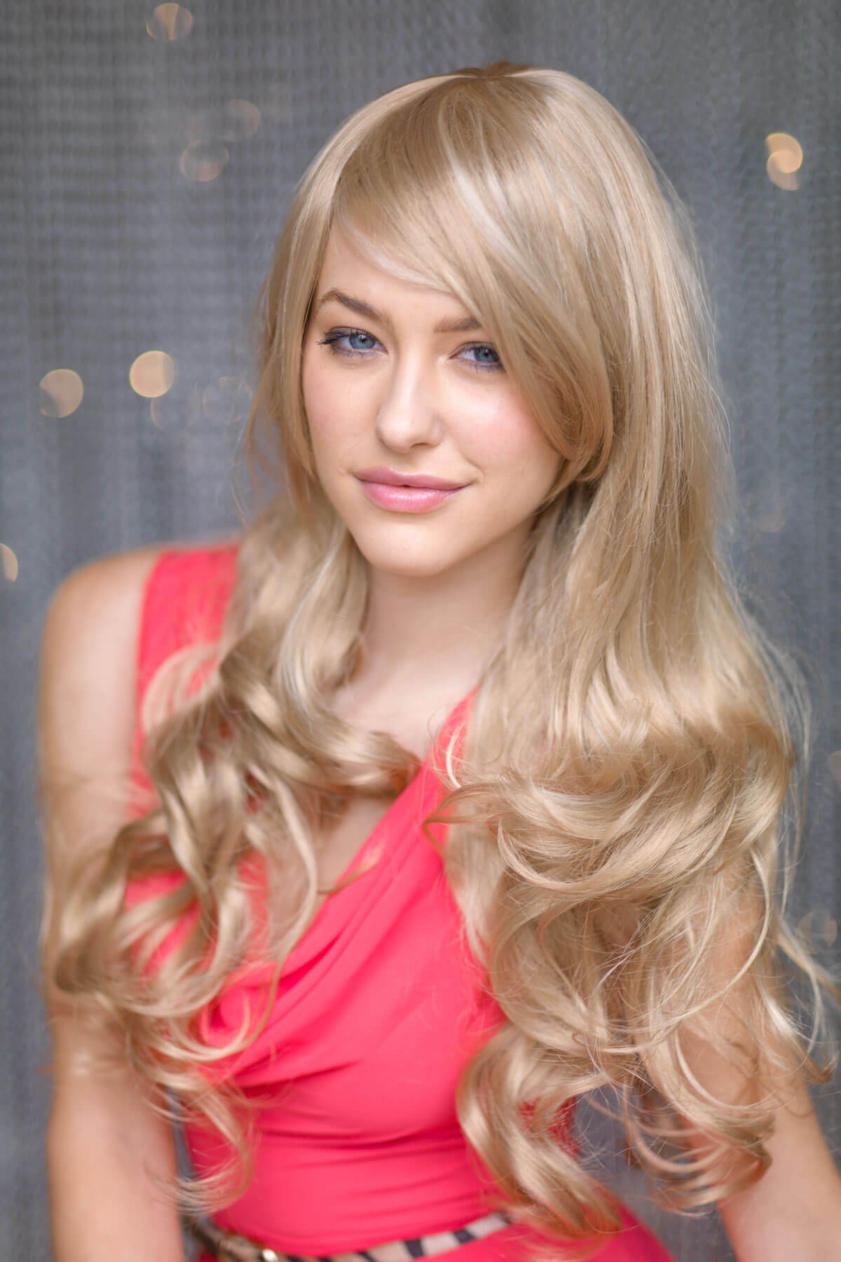 Woman with long blonde hair wearing a red top against a gray background. Blonde wig with big loose curls and sweeping fringe, extra long: Carly ash blonde and light blonde 24H613