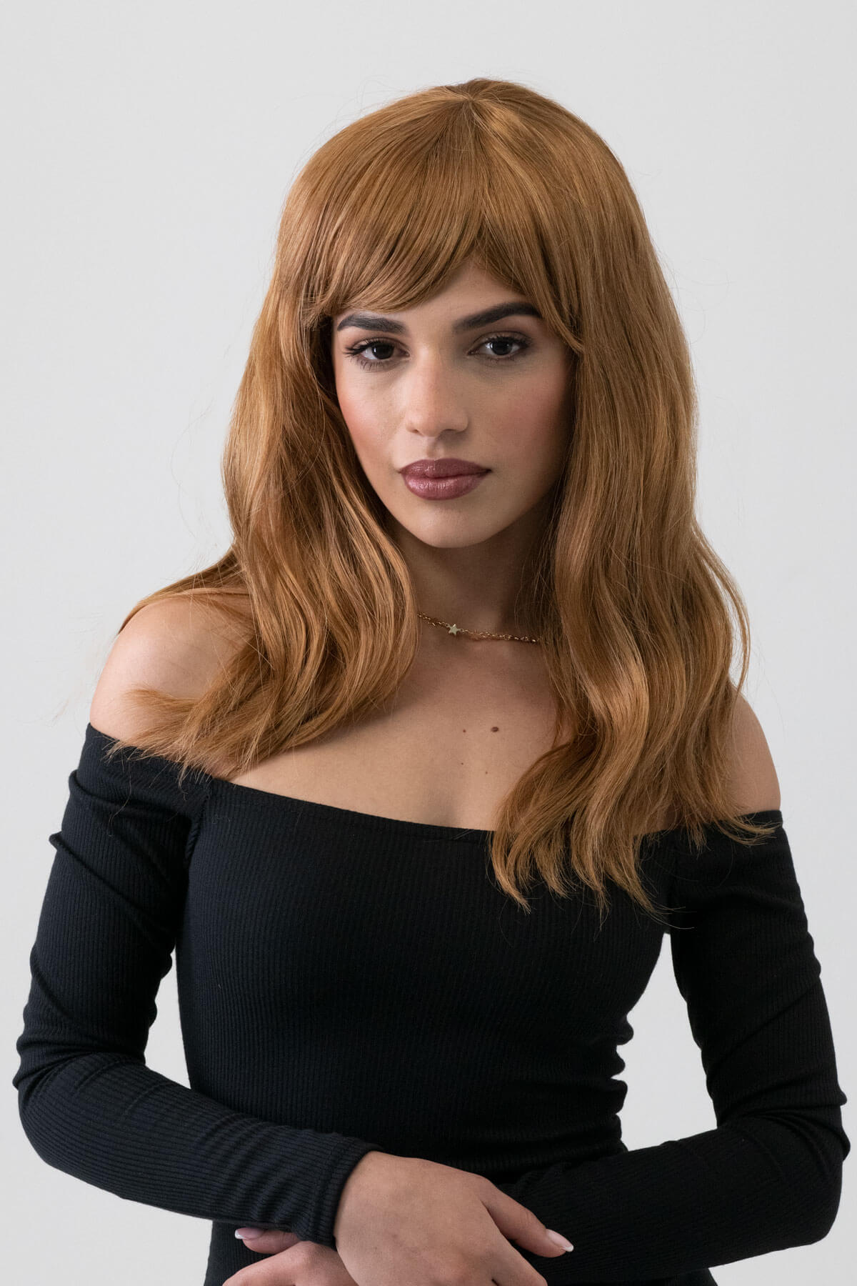 Woman with a brown wig wearing a black off-shoulder top against a white background. Golden brown wig with long waves: Carmella