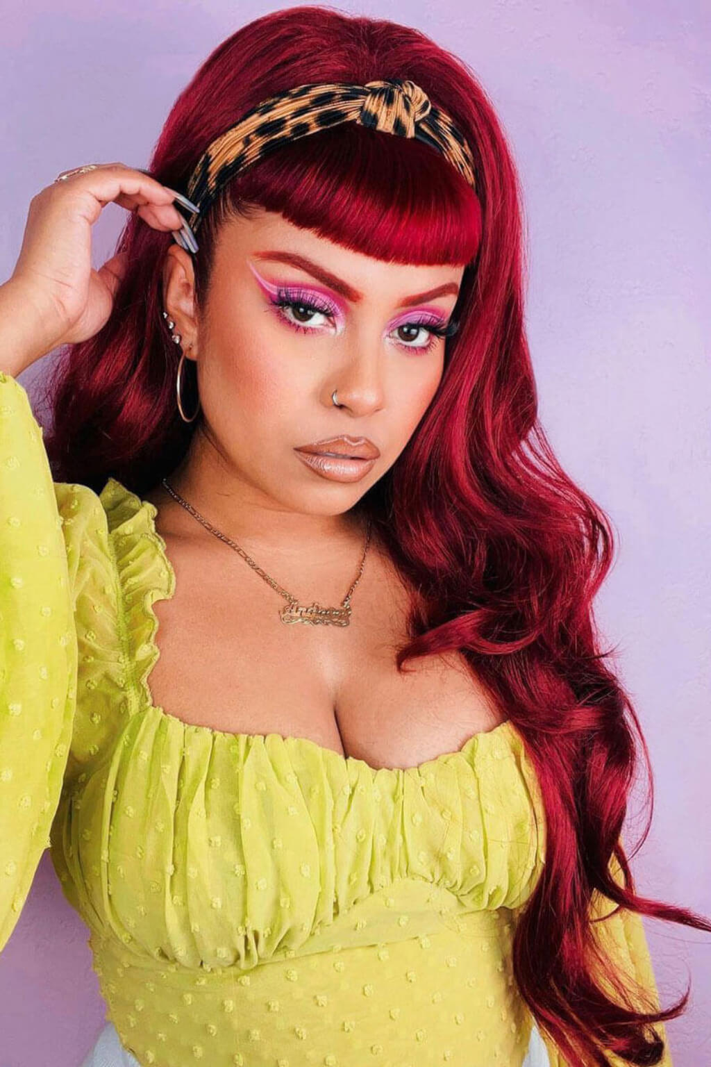 Woman with vibrant red hair and colorful makeup wearing a yellow top against a light purple background. High crown long, wavy half wig and volume ring: Bardot brown and blonde 6H24