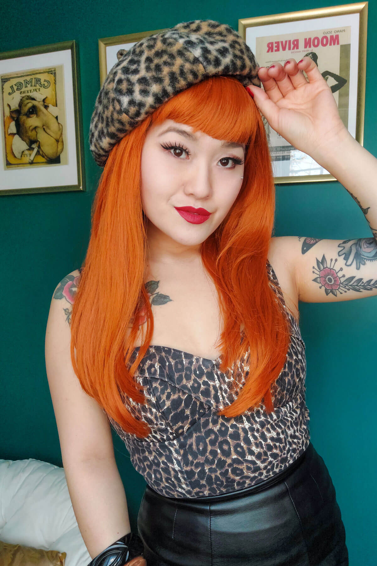 Person with orange hair and leopard print outfit against a teal wall with framed pictures. Long ginger wig with straight hair and a short, straight fringe: Daphne